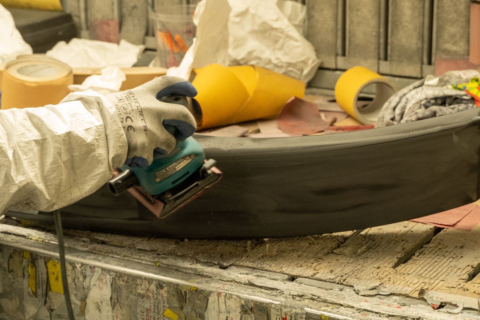 Una mano guantata carteggia un pannello grigio di un'auto in un'officina, con attrezzi e materiali di mascheratura nelle vicinanze.