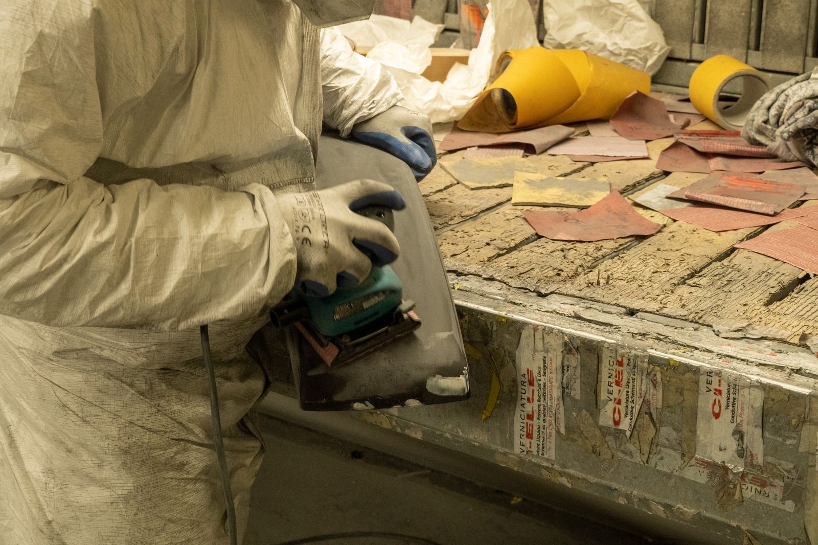 Un operaio con una tuta bianca utilizza una levigatrice manuale su una superficie di cemento scheggiata, con delle piastrelle nelle vicinanze.