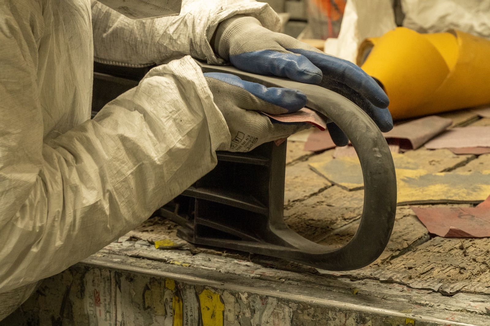 Un operaio con i guanti tiene in mano un attrezzo ad anello in metallo nero sopra un banco da lavoro polveroso con piastrelle e indossa un elmetto giallo.