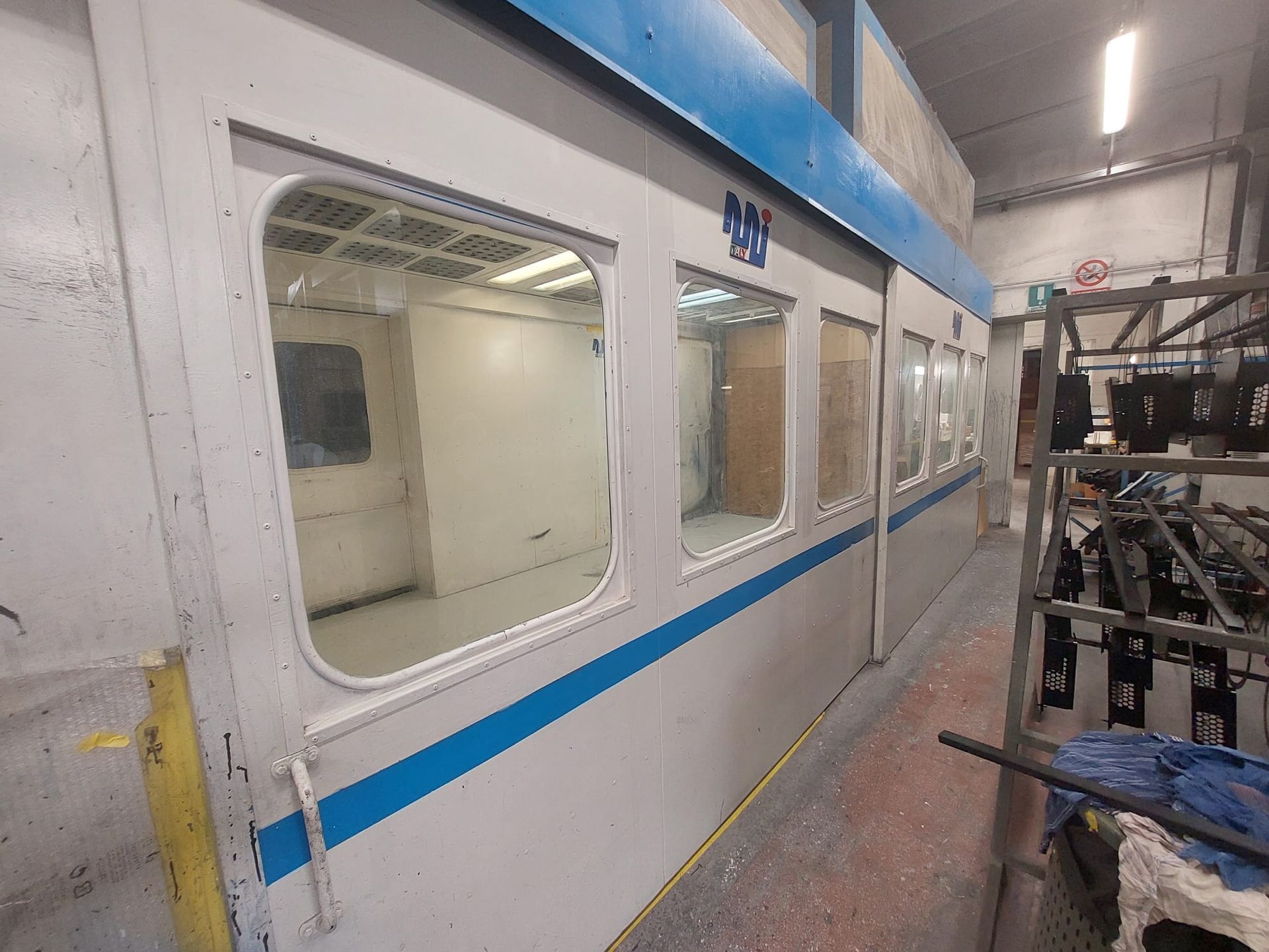 Cabina di verniciatura industriale con pareti bianche, finiture blu e finestre all'interno di un ambiente di lavoro simile a una fabbrica.