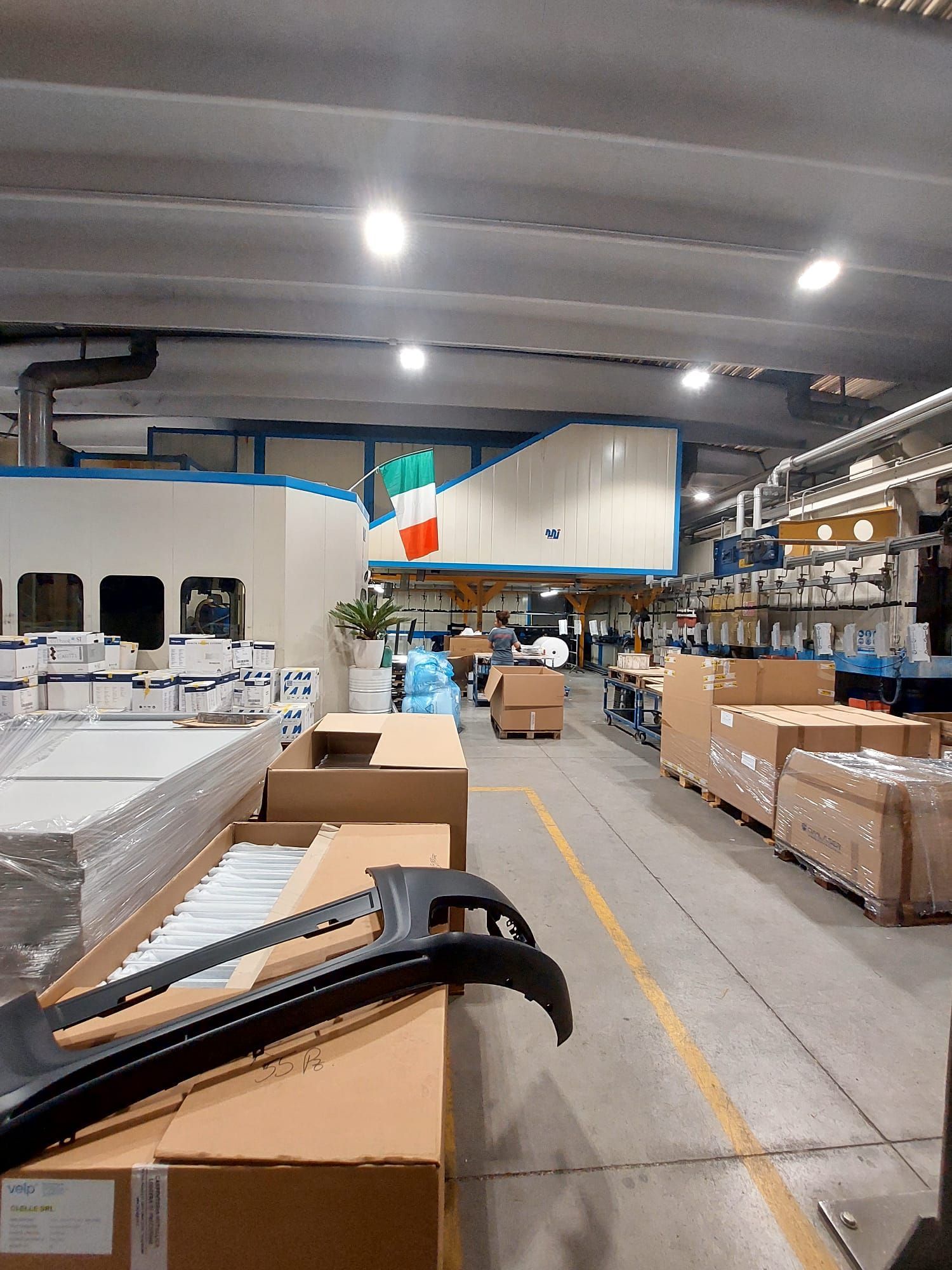 Corridoio di magazzino con scatole impilate, attrezzature industriali e luci a soffitto.