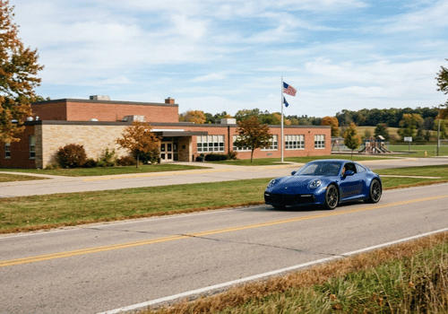 Blue Porsche 911 driving past a school on a sunny early fall day, highlighting back-to-school