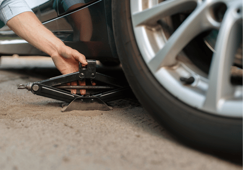 Hand using a jack to lift a silver car’s wheel on a roadside