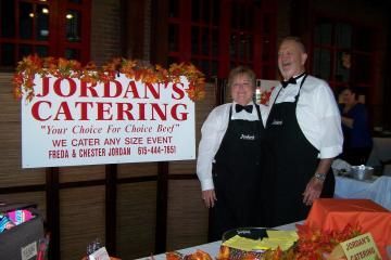 Two people in black aprons stand beside a Jordan’s Catering sign at an indoor event.
