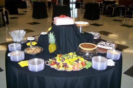Dessert buffet table with assorted cakes, fruit, and covered trays on black tablecloths in a banquet hall