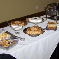 Dessert buffet table with pies, pastries, cookies, and a coffee urn on a white tablecloth.