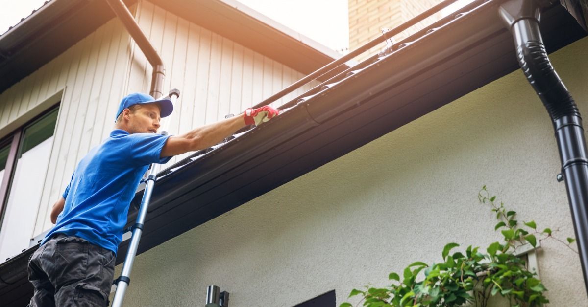 Upward view of a worker touching the top of a house's roof gutter with gloved hands. He stands on a ladder.