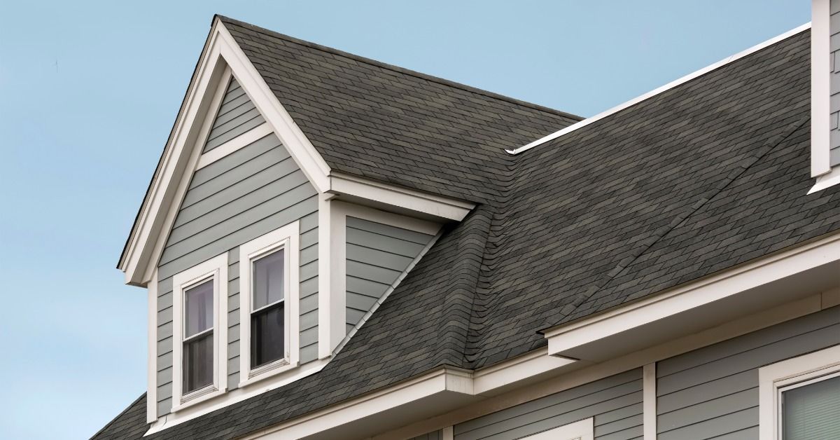 Upward view of the roof of a gray house with white trim. A dormer with two windows extends from it.