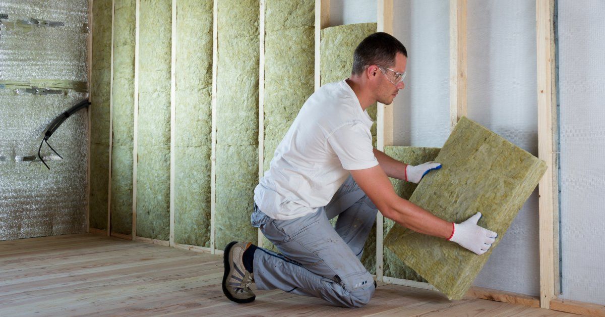 A worker kneels while placing a thick insulation batt between the wooden studs of an unfinished wall. He wears gloves.