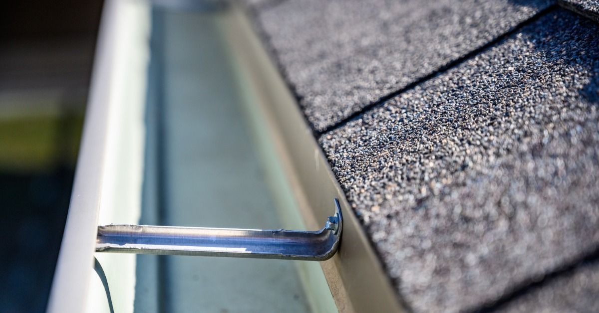 A cleaned-out gutter runs beside textured roof shingles. Metal gutter hangers connect the gutter and the roof edge.