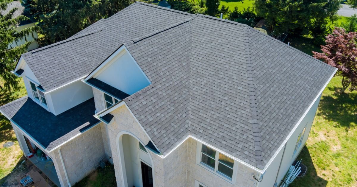 Aerial view of a home with asphalt shingles and multiple roof slopes. Trees, lawn, and a driveway.