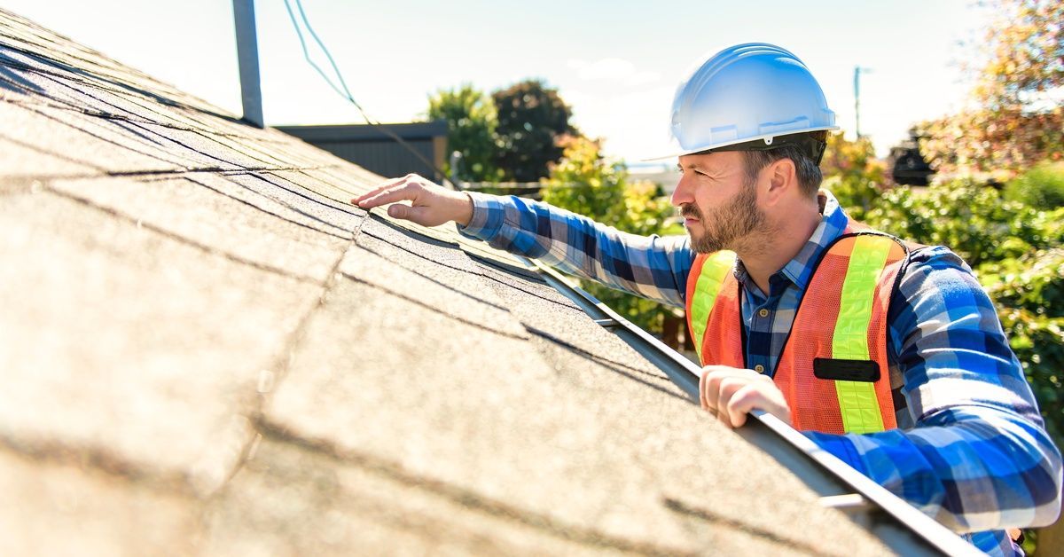 A roof inspector wearing a hard hat and safety vest touches and examines asphalt shingles on a slope