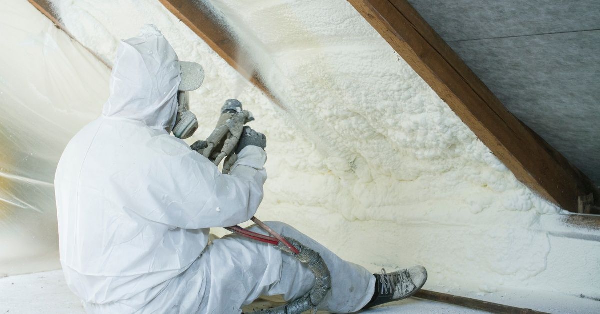 A worker in a white protective suit sits under sloped roof framing and applies spray foam insulation between the beams.