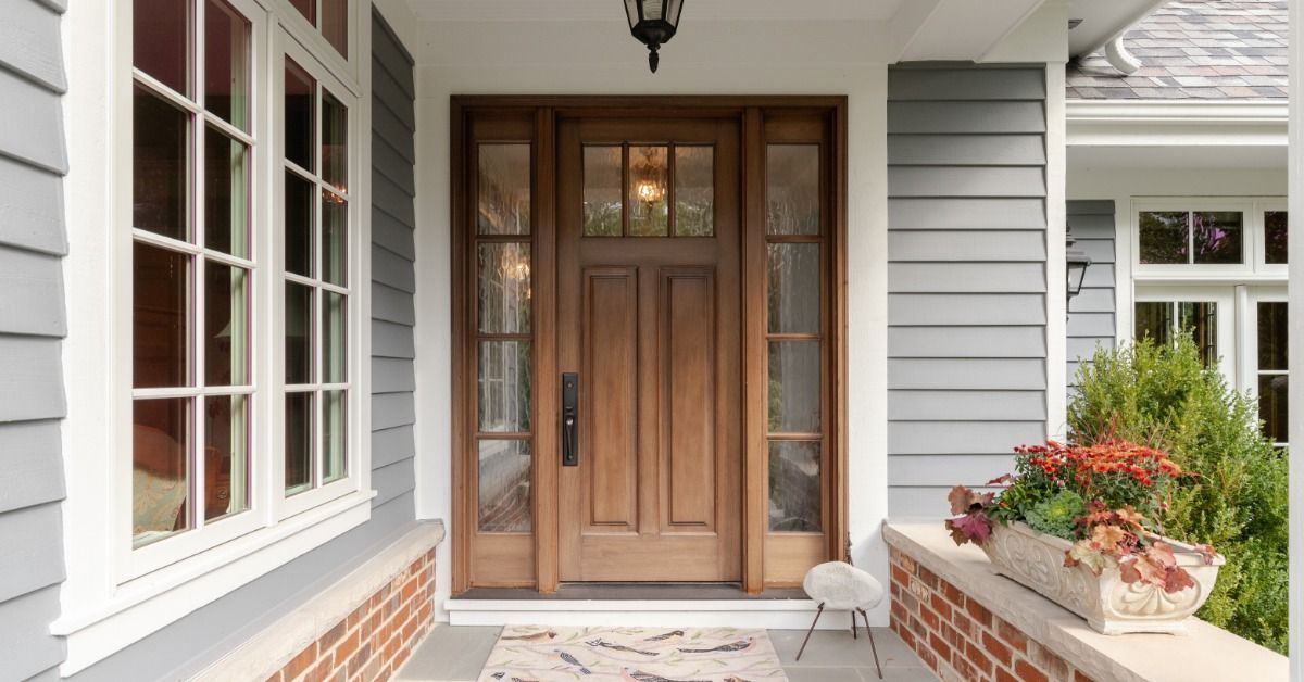 A wooden front door with glass side panels is beneath a covered porch. White-trimmed windows line the adjacent wall.