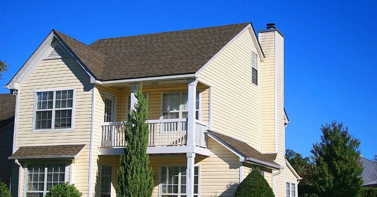A pale yellow two-story house has horizontal siding, white trim, and a covered upper balcony. Shrubs surround the house.