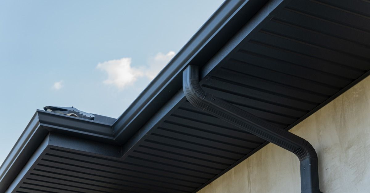 Upward view of black gutters, a black downspout, and black soffit panels above a light cream-colored wall.