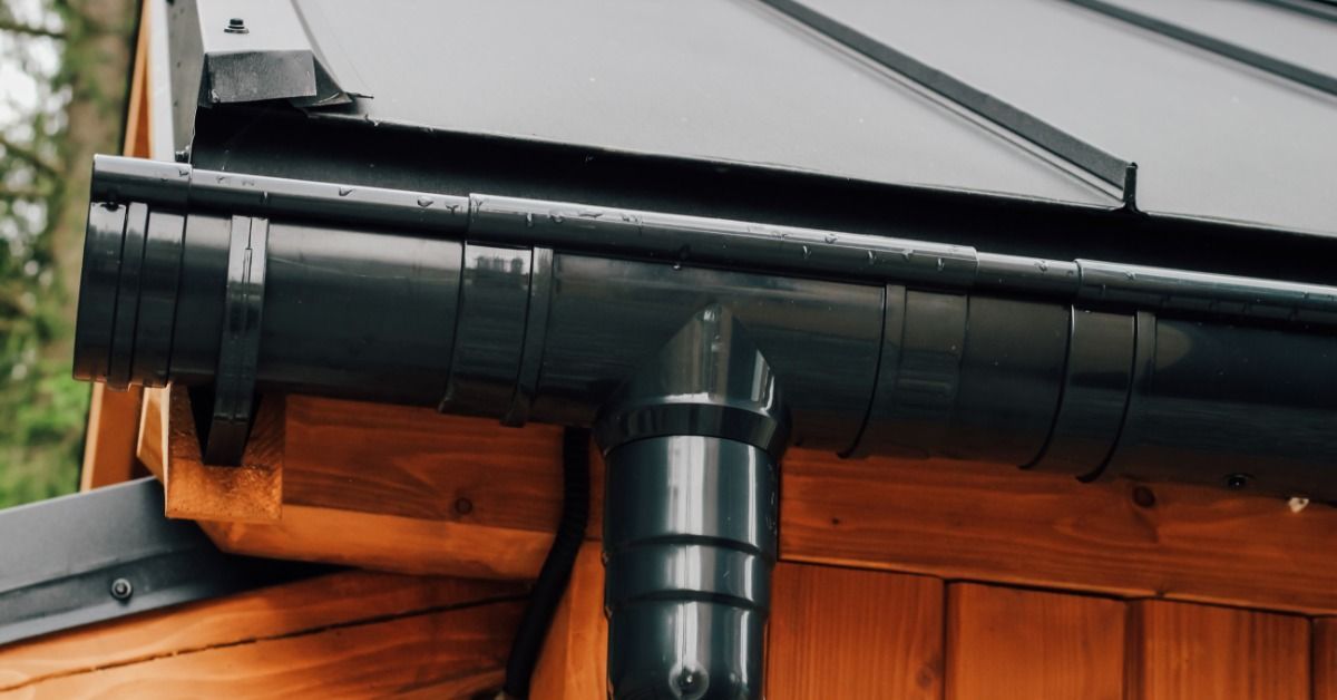A black, half-round gutter runs along the edge of a roof on a wood-sided structure. A downspout connects to the gutter.