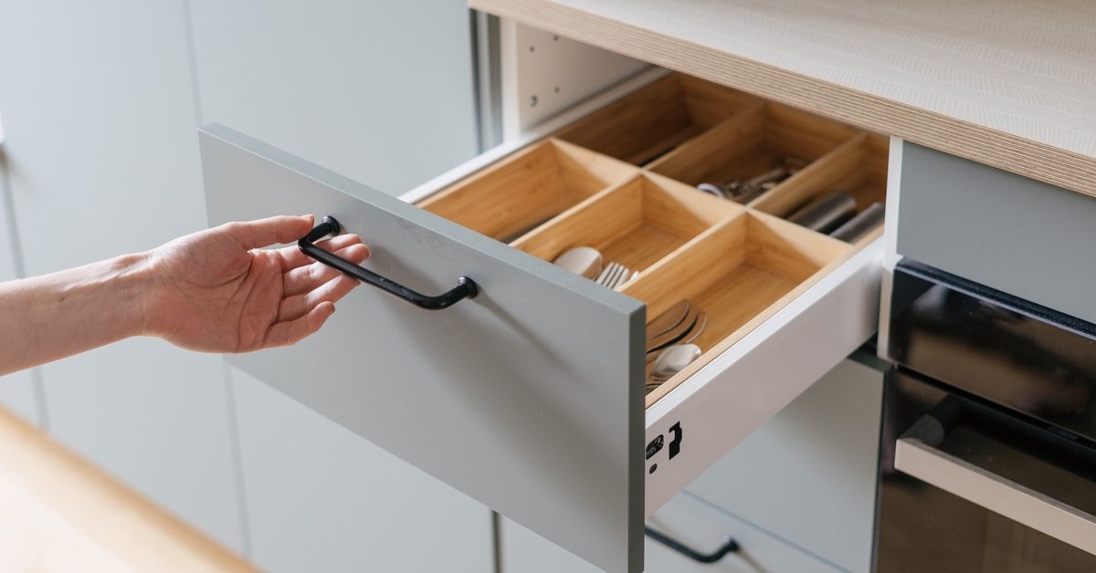 A hand pulls open a gray kitchen drawer with a black metal handle. A wooden organizer inside separates utensils.