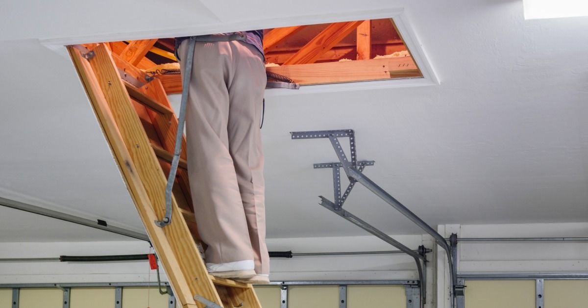 A person stands on an attic ladder leading to an open ceiling hatch in a garage. Wooden framing is visible in the attic.