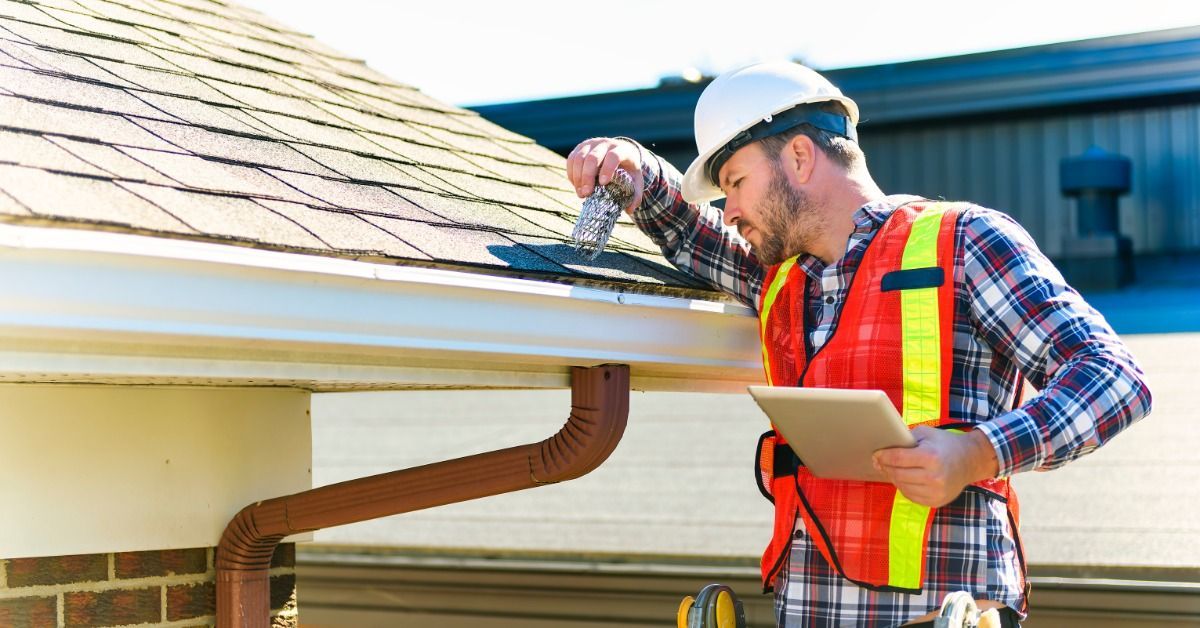 An inspector in a hard hat and reflective vest stands on a ladder beside a shingled roof.