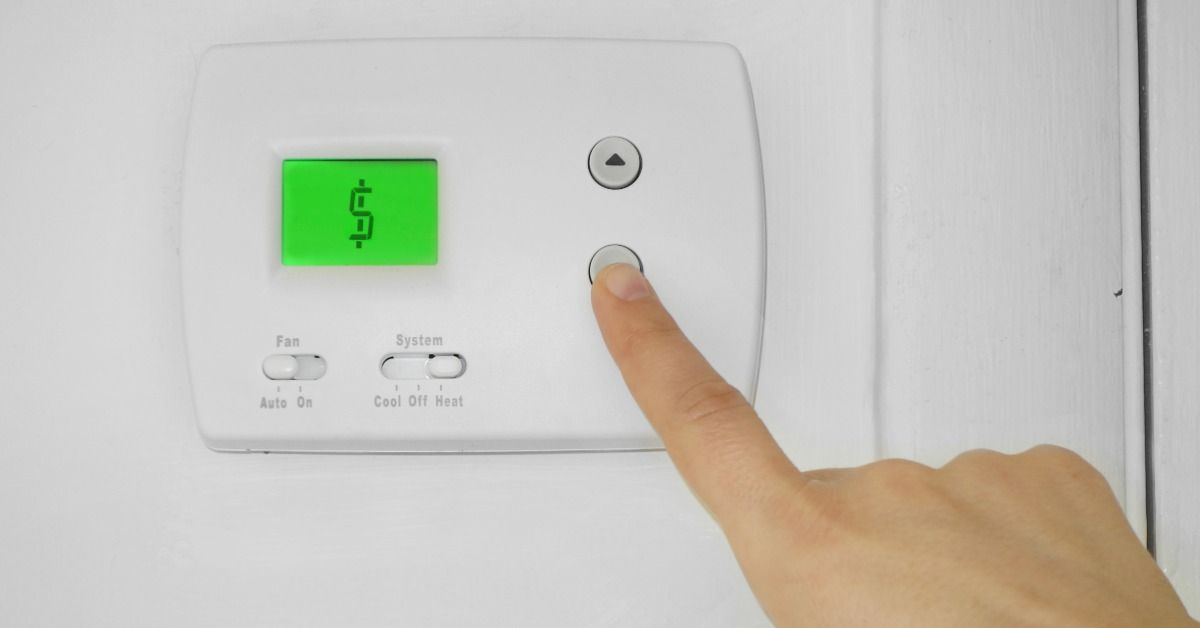 A person presses the down arrow button on a wall-mounted thermostat. The green display screen shows a dollar sign.