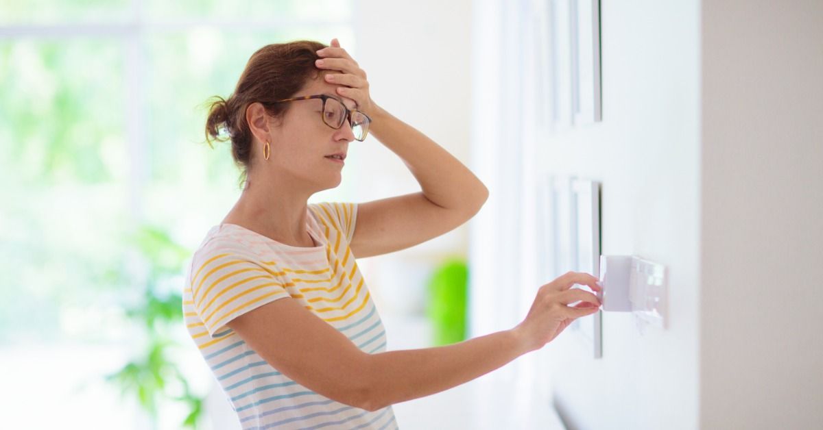 A woman in a bright room holds a hand to her forehead as she adjusts a thermostat.
