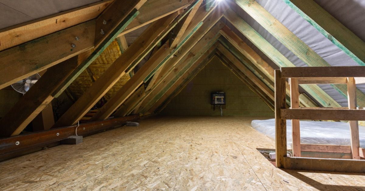 An unfinished attic has exposed wooden rafters, roof framing, and a plywood floor. A bright light shines from the roof.
