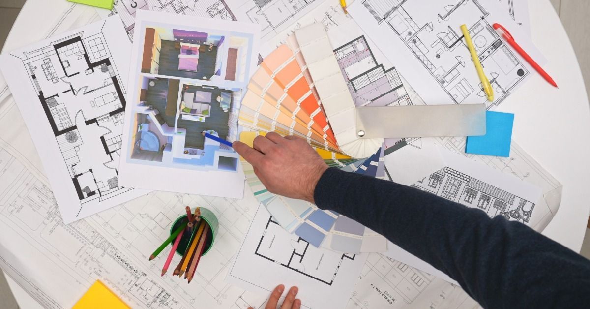 Architectural floor plans and paint color samples are on a table. A person points to an interior design rendering.