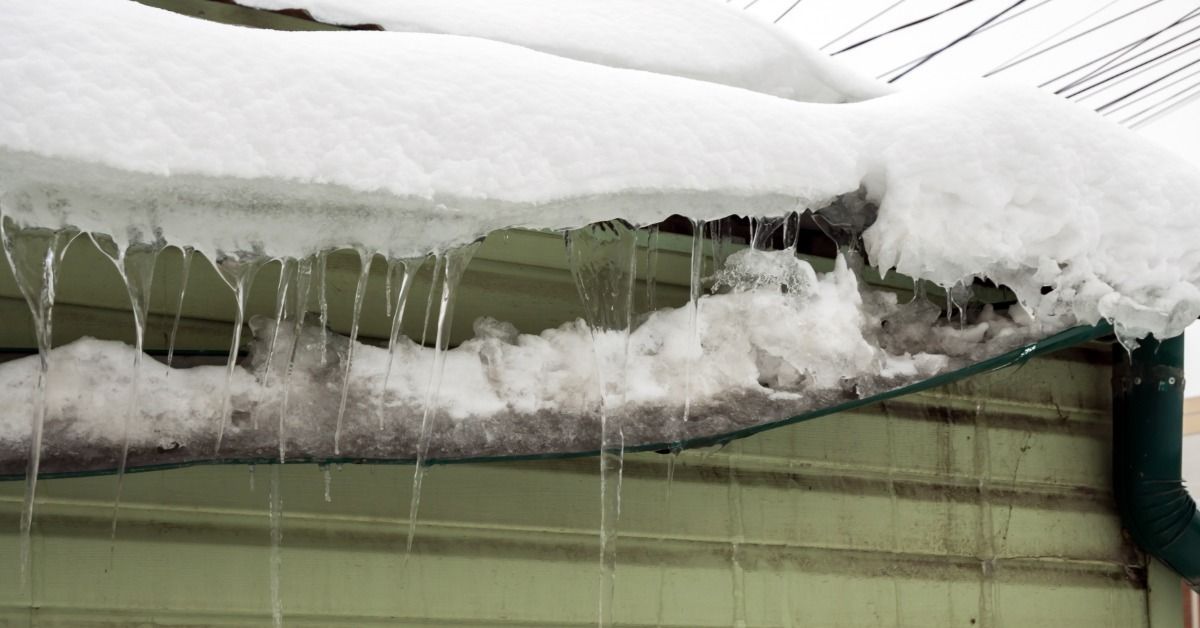 A snow-covered gutter hangs down along the edge of a house. Several icicles hang from the gutter and roofline.
