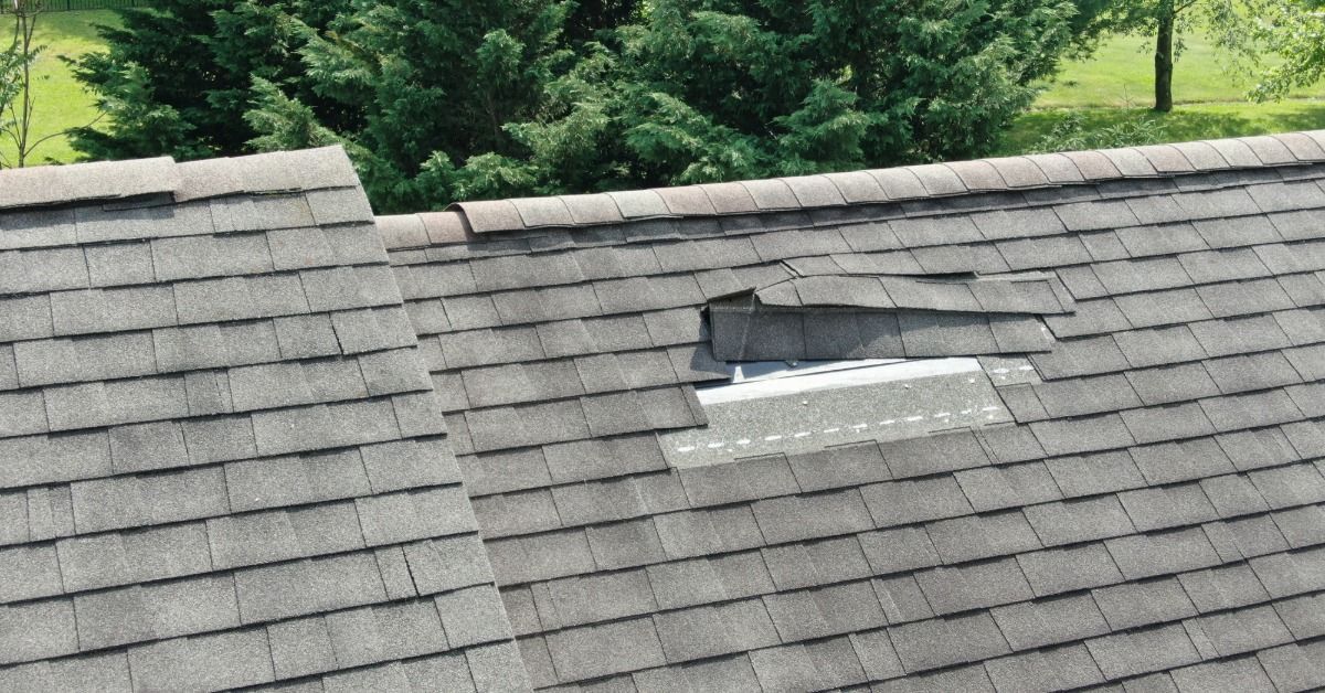 Gray asphalt shingles cover a sloped roof, but some shingles are missing near the center. Trees are behind the roof.