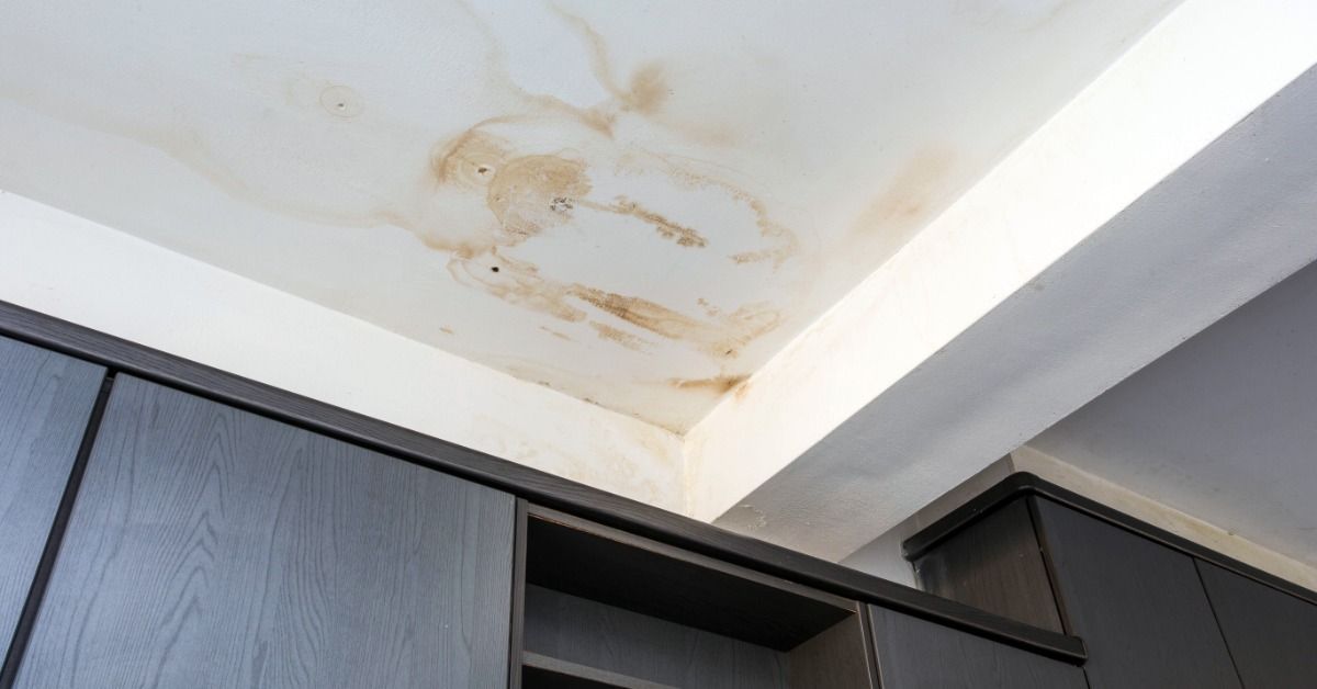 A white ceiling has several brown water marks and streaks above a row of dark cabinets. The stains extend from a corner.