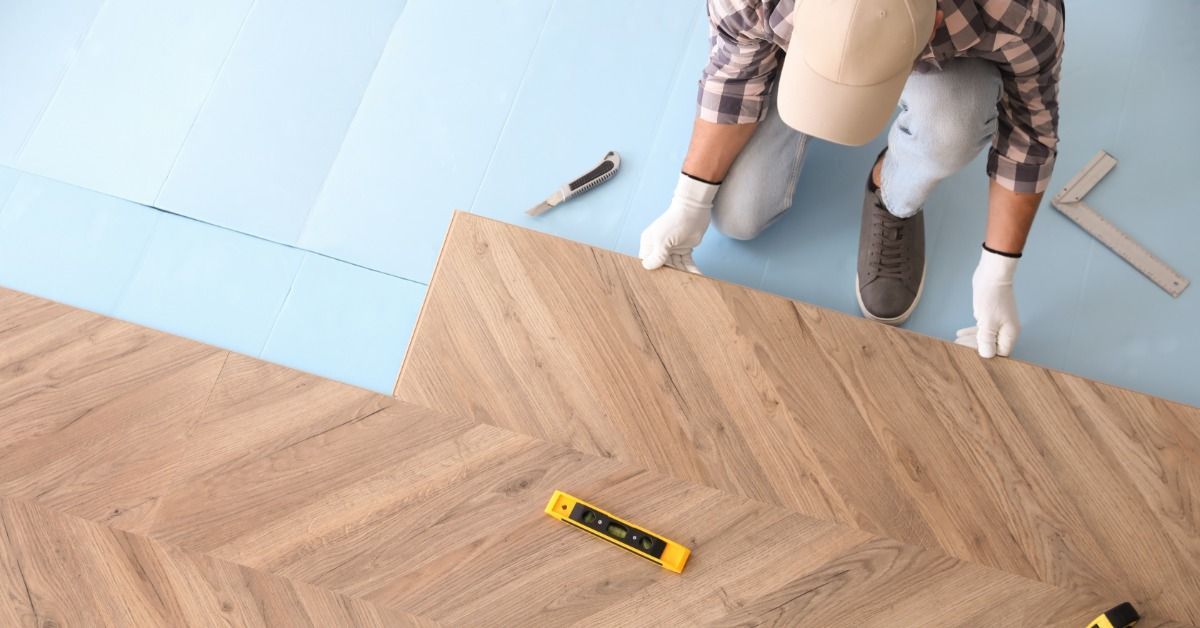 A worker positions a flooring plank over blue underlayment. A tape measure and a level are nearby.