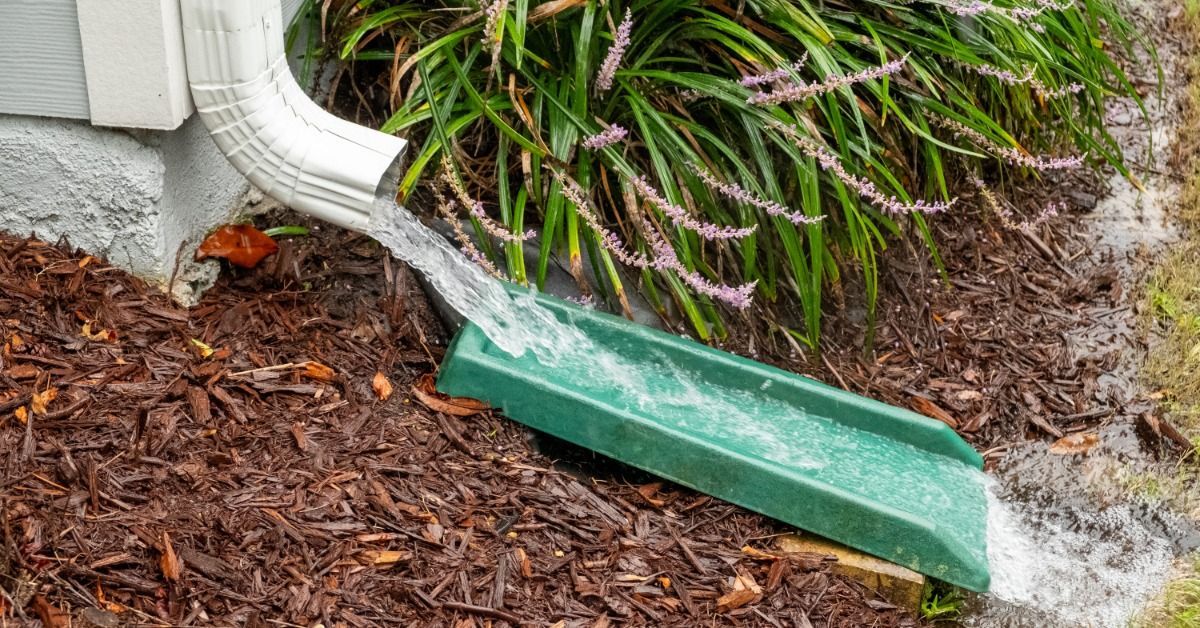 Water flows from a white downspout onto a green splash block by a home's foundation. Mulch surrounds the drainage area.