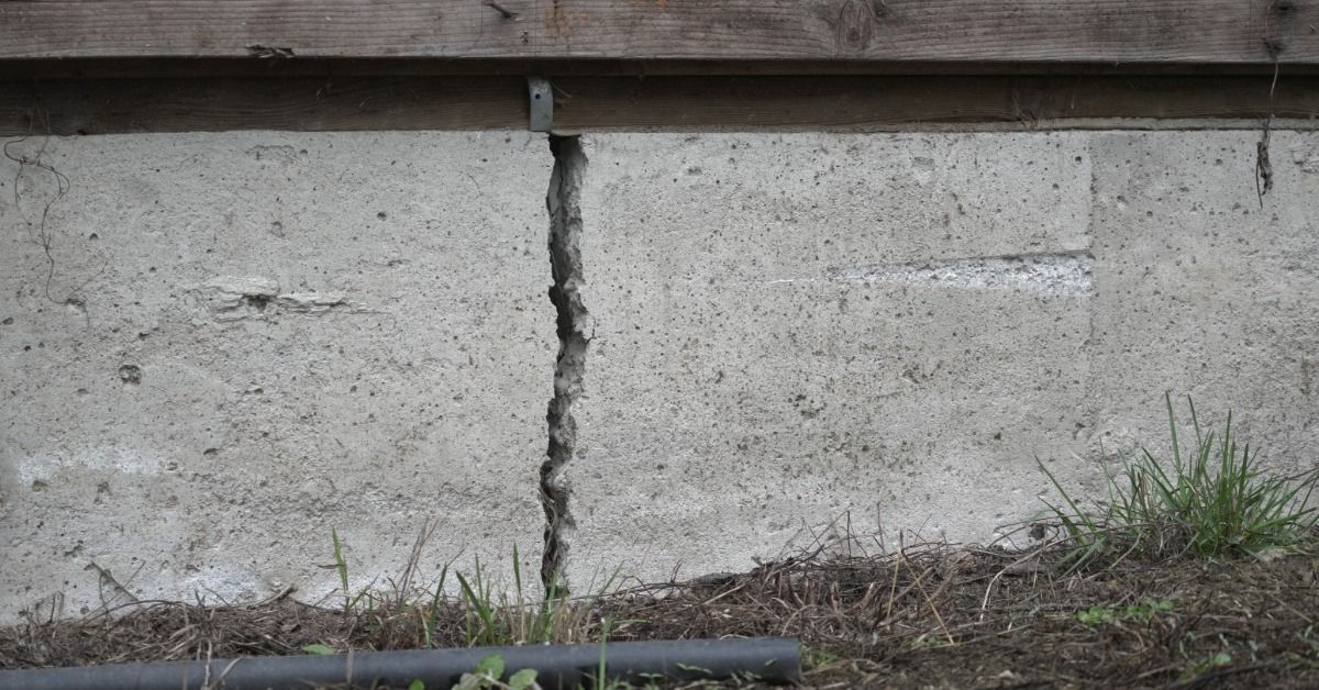 A crack goes from the top of a concrete foundation to the ground. A wooden board runs horizontally above the foundation.
