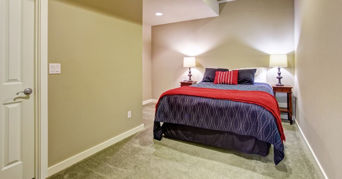 A carpeted bedroom has exposed, boxed-in beams, neutral-colored walls, and a large bed with two bedside lamps.