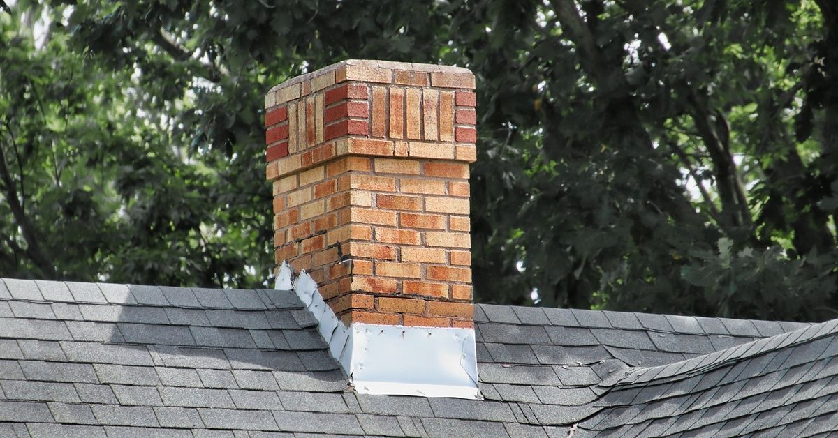 A brick chimney with metal flashing at its base tops an asphalt shingle roof. Leafy tree branches fill the background.