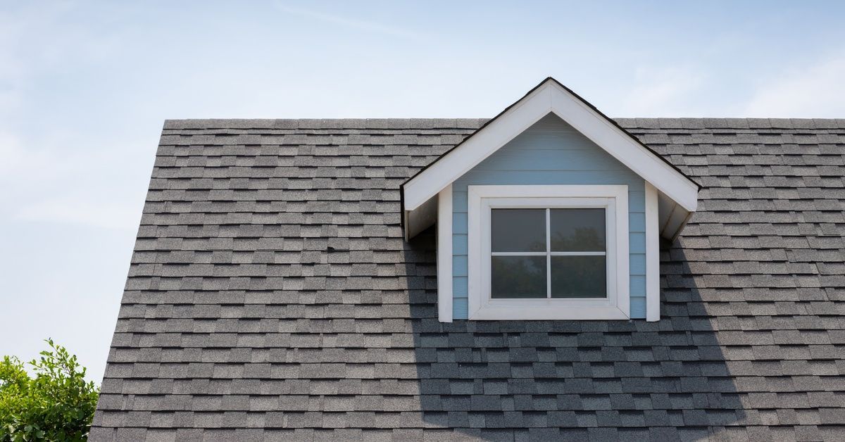An asphalt shingle roof slopes downward and surrounds a blue dormer with white trim and a window.