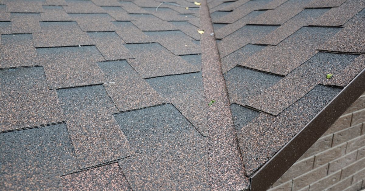 Two slopes of a shingled roof meet at a valley lined with the same roofing material. A few leaves are on the roof.