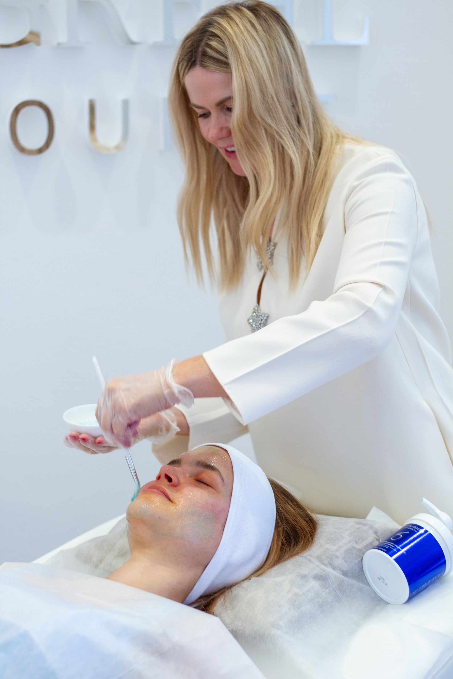 Eine Kosmetikerin führt eine Gesichtsbehandlung an einer Kundin durch, die mit einem Stirnband auf einer Spa-Liege liegt.