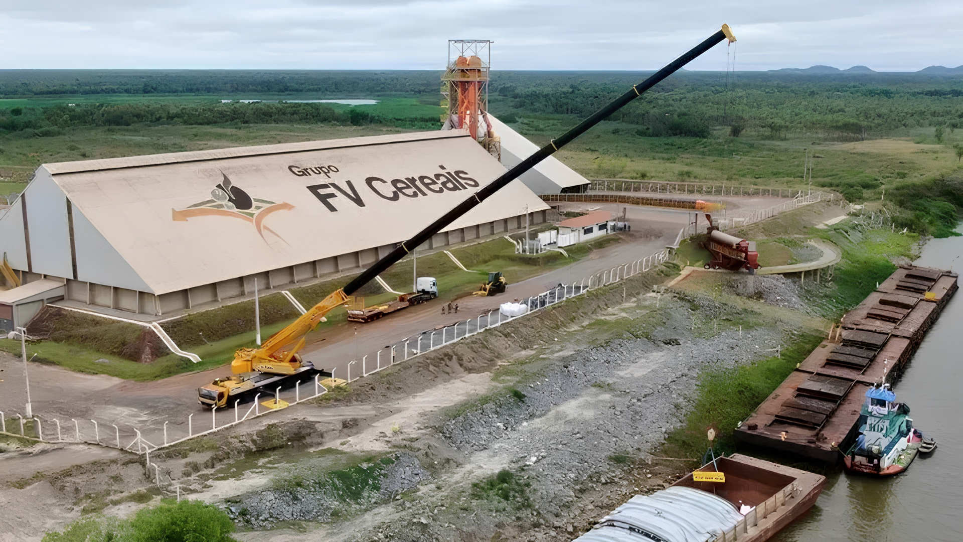 Vista aérea de um grande galpão industrial branco, guindastes, caminhões e um canal ao lado