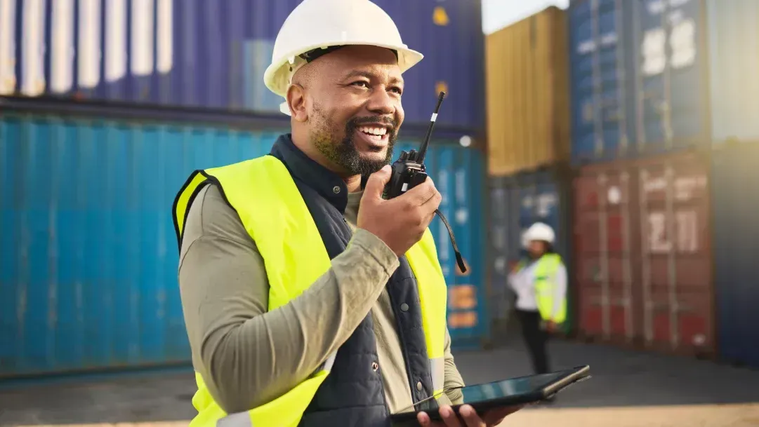 Um homem usando capacete e colete de segurança está falando em um walkie talkie enquanto segura um tablet.