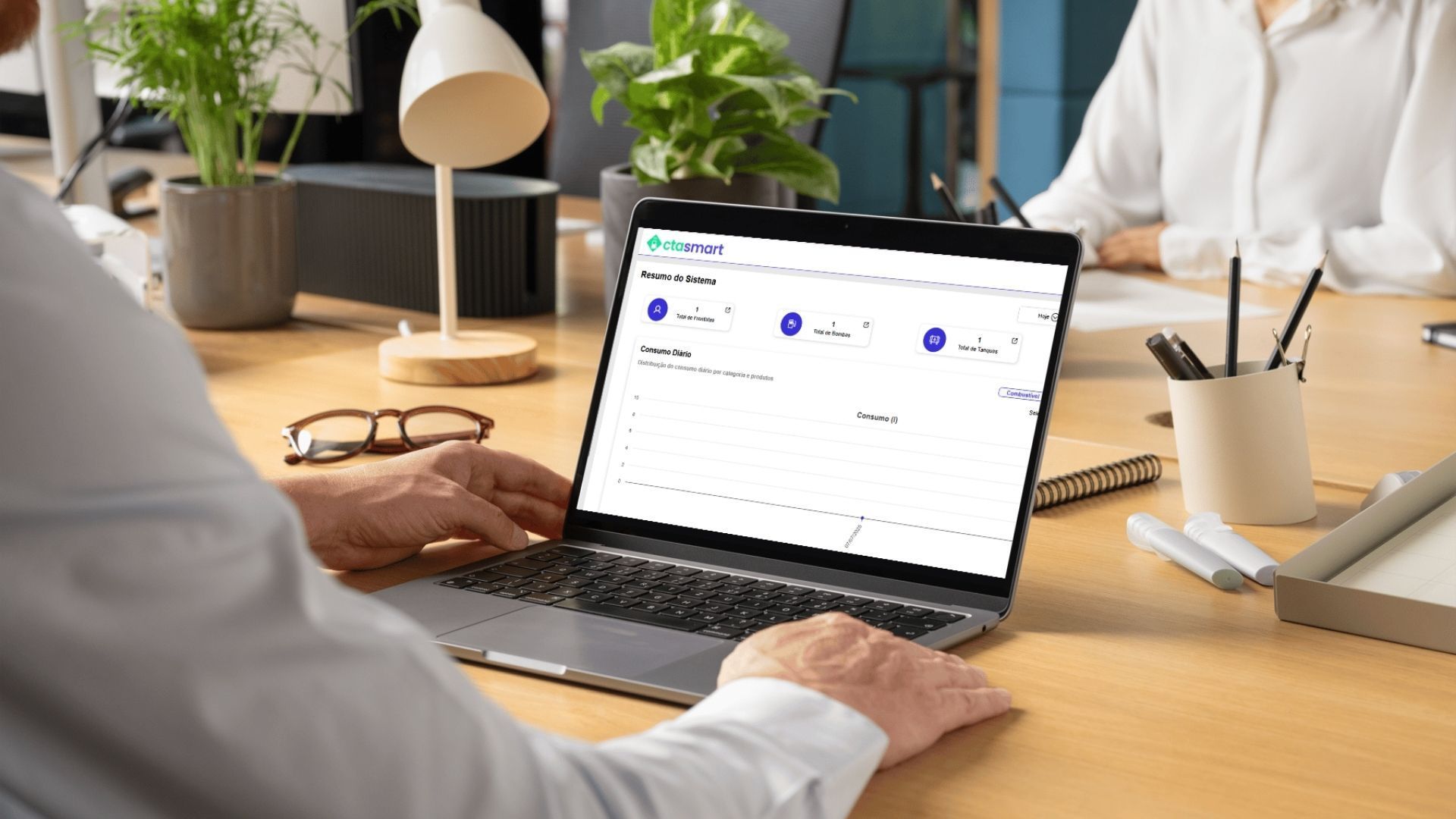 Homem usando um laptop em uma mesa, analisando dados. Outra pessoa está ao fundo. Ambiente de escritório.
