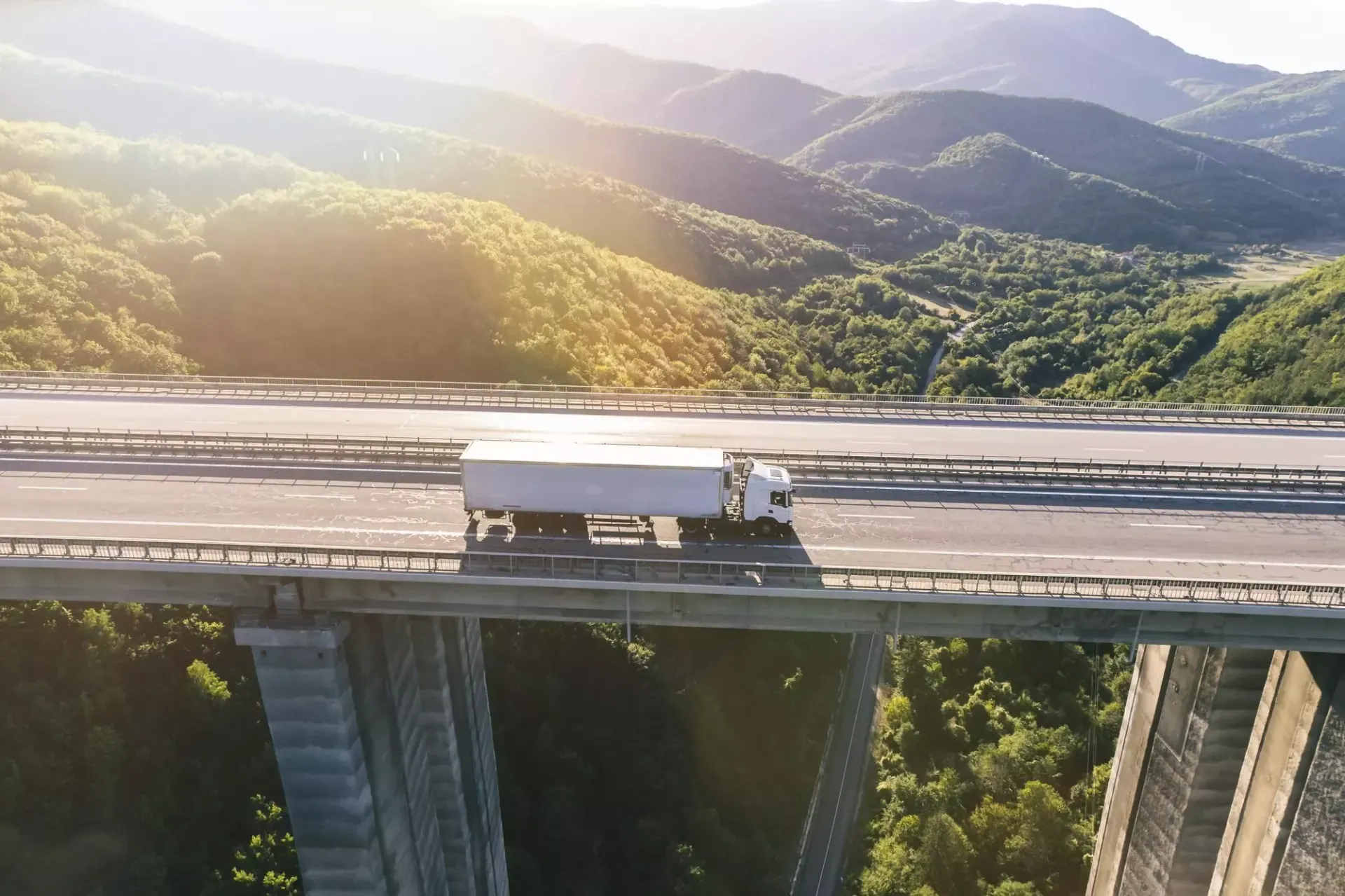 Caminhão atravessando uma ponte alta com montanhas e luz solar ao fundo.