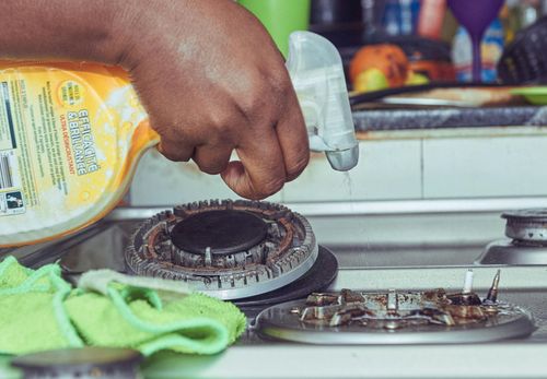 Hand spraying cleaner on a dirty stovetop burner. A green cloth sits nearby.