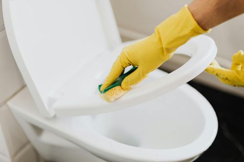 Person wearing yellow gloves cleans a white toilet seat with a green sponge.