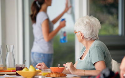 Woman painting a wall while an older person watches at a table with fruit indoors