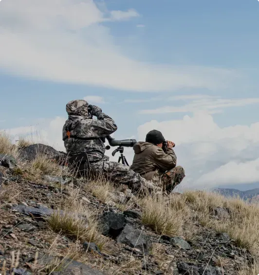 Two soldiers are sitting on top of a hill looking through binoculars.