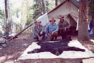 Three men are kneeling next to a bear rug in front of a tent.