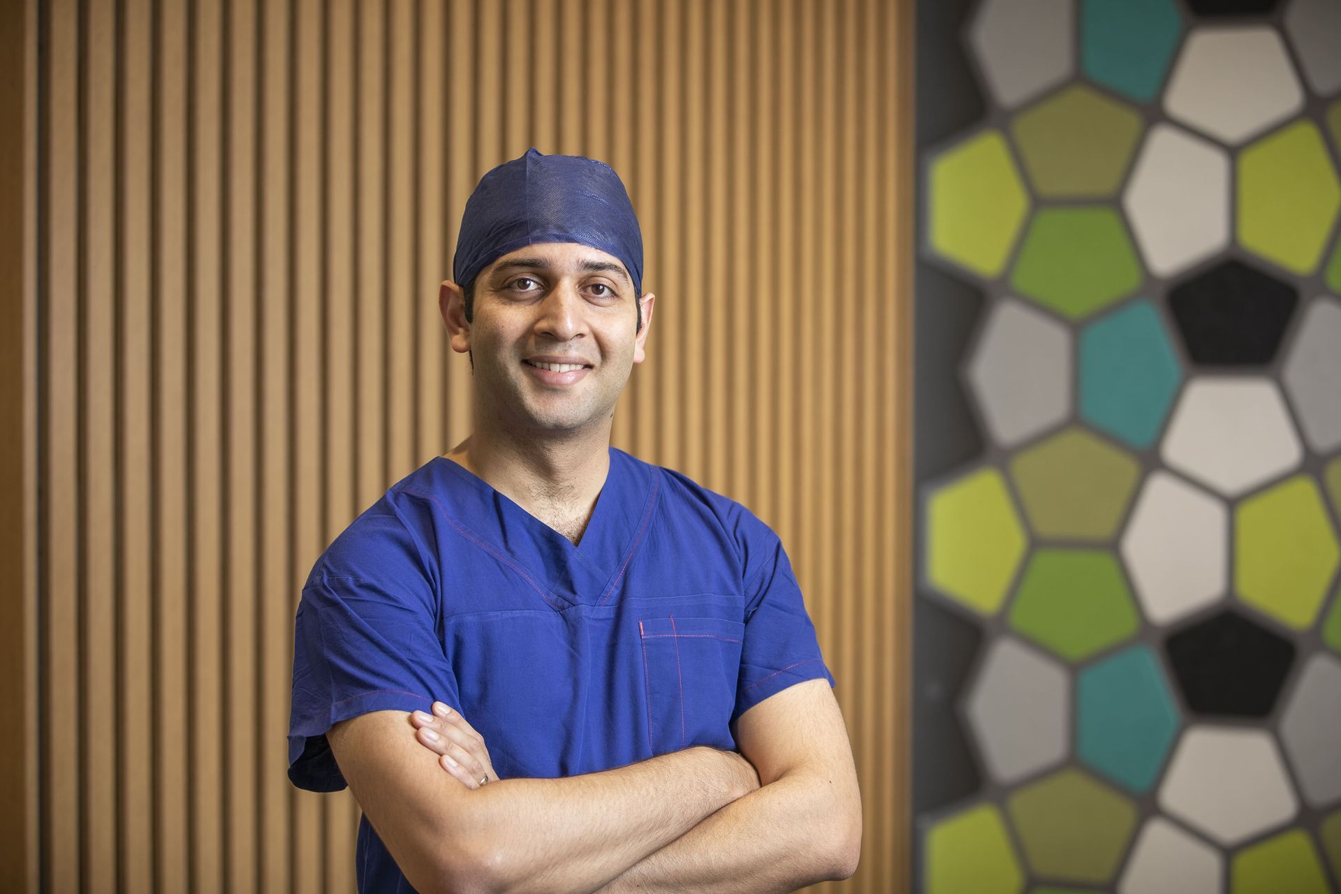 Smiling medical professional in blue scrubs with arms crossed, standing before a wood and geometric-pattern wall.