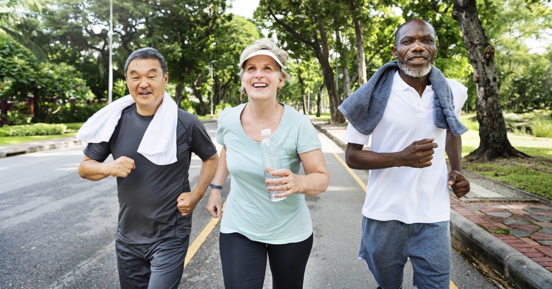Three friends jogging on a tree-lined road, smiling and wearing casual athletic clothes.